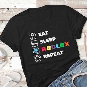 Puede incluir: Una camiseta negra de manga corta con un estampado gr&aacute;fico blanco con el texto EAT, SLEEP, ROBLOX, REPEAT. Cada palabra va acompa&ntilde;ada de un icono cuadrado, y la palabra ROBLOX se muestra en letras multicolores que incluyen rojo, amarillo, azul y naranja.