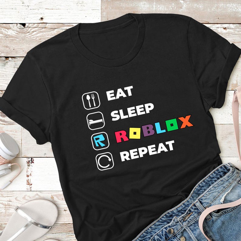 Puede incluir: Una camiseta negra de manga corta con un estampado gr&aacute;fico blanco con el texto EAT, SLEEP, ROBLOX, REPEAT. Cada palabra va acompa&ntilde;ada de un icono cuadrado, y la palabra ROBLOX se muestra en letras multicolores que incluyen rojo, amarillo, azul y naranja.