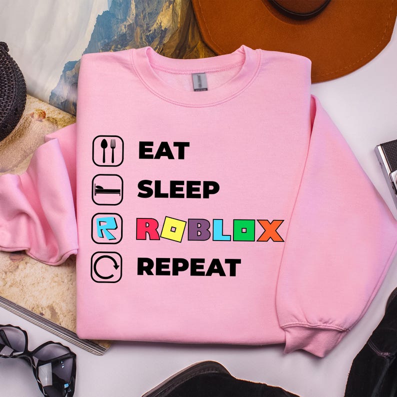 Puede incluir: Una sudadera de manga larga de color rosa claro con un estampado gr&aacute;fico con el texto EAT SLEEP ROBLOX REPEAT. El dise&ntilde;o incluye iconos de una cuchara y un tenedor, una cama, la letra R en un cuadrado azul y una flecha circular con letras.