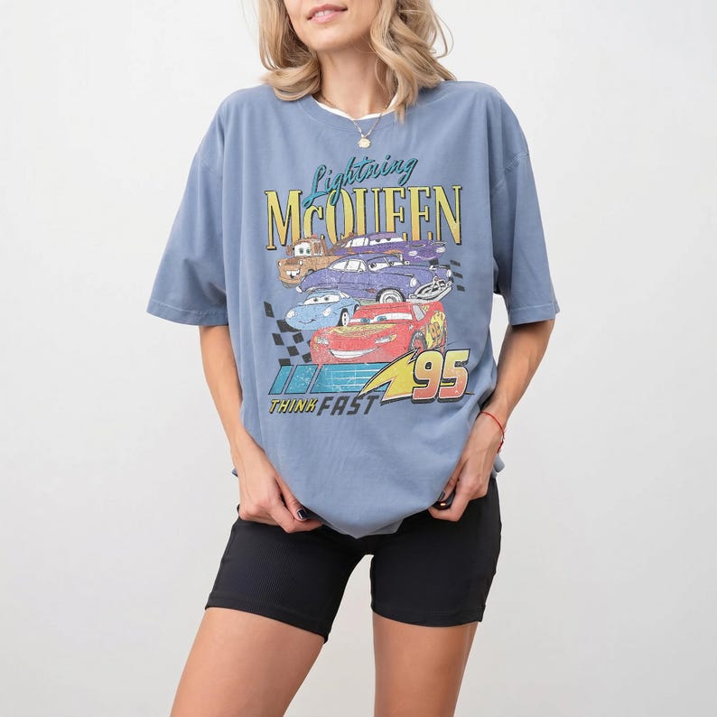 Puede incluir: Camiseta azul claro con un gr&aacute;fico de Rayo McQueen y otros personajes de Cars. El texto "Lightning McQueen" y "95" son visibles. La camiseta tiene cuello redondo y mangas cortas.