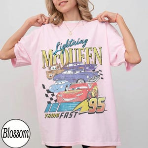 Puede incluir: Camiseta rosa claro con un dise&ntilde;o de Rayo McQueen y otros personajes de Cars. El dise&ntilde;o incluye el texto "Lightning McQueen", "95" y "Think Fast". La camiseta tiene mangas cortas y un corte holgado.
