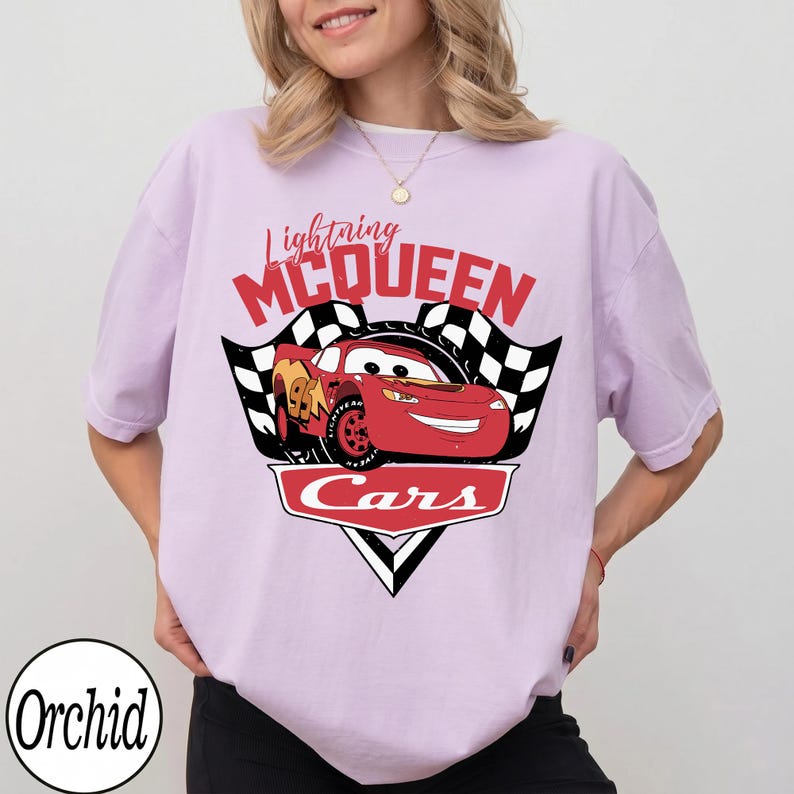 May include: A light purple t-shirt featuring a graphic of Lightning McQueen from the movie Cars. The design includes the text "Lightning McQueen" in red, a red race car, and checkered flags. The shirt has short sleeves.