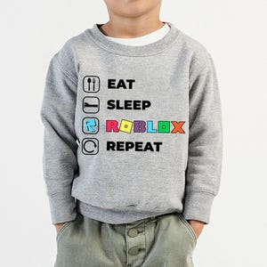 Puede incluir: Una sudadera gris jaspeado de manga larga con un estampado gr&aacute;fico que incluye el texto EAT, SLEEP, ROBLOX y REPEAT. Cada palabra va acompa&ntilde;ada de un icono correspondiente: cuchara y tenedor, una cama, el logotipo de Roblox y una flecha.