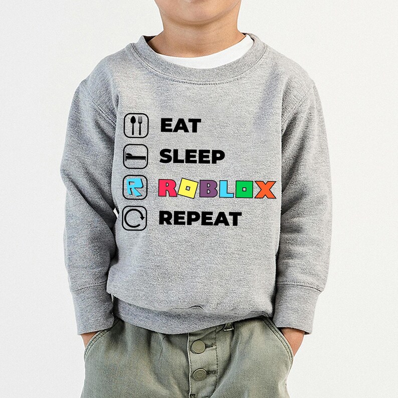 Puede incluir: Una sudadera gris jaspeado de manga larga con un estampado gr&aacute;fico que incluye el texto EAT, SLEEP, ROBLOX y REPEAT. Cada palabra va acompa&ntilde;ada de un icono correspondiente: cuchara y tenedor, una cama, el logotipo de Roblox y una flecha.