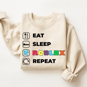 Puede incluir: Una sudadera de cuello redondo color beige con un estampado gr&aacute;fico de cuatro filas. El texto dice EAT, SLEEP, ROBLOX y REPEAT en negro, con la palabra ROBLOX en letras de colores. Cada l&iacute;nea incluye un icono negro: cubiertos, cama, logo y flecha.