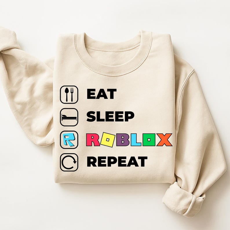 Puede incluir: Una sudadera de cuello redondo color beige con un estampado gr&aacute;fico de cuatro filas. El texto dice EAT, SLEEP, ROBLOX y REPEAT en negro, con la palabra ROBLOX en letras de colores. Cada l&iacute;nea incluye un icono negro: cubiertos, cama, logo y flecha.