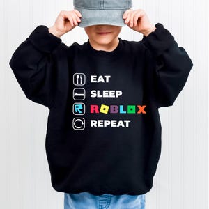Puede incluir: Una sudadera negra de manga larga con texto e iconos en blanco. El dise&ntilde;o muestra las palabras EAT, SLEEP, ROBLOX y REPEAT, cada una acompa&ntilde;ada de un icono correspondiente: una cuchara y un tenedor, una cama, el logotipo de Roblox y una flecha circular.