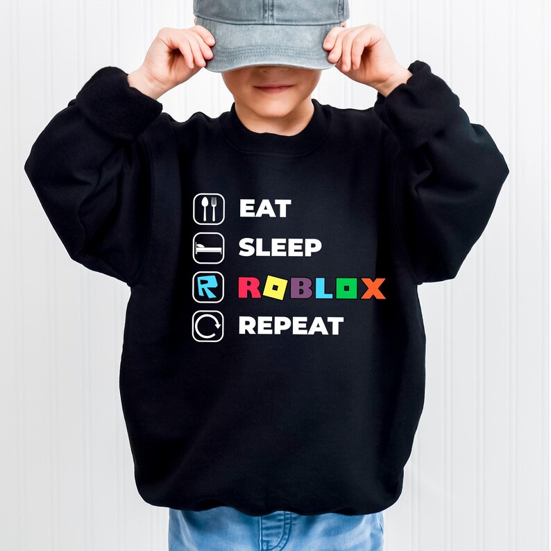Puede incluir: Una sudadera negra de manga larga con texto e iconos en blanco. El dise&ntilde;o muestra las palabras EAT, SLEEP, ROBLOX y REPEAT, cada una acompa&ntilde;ada de un icono correspondiente: una cuchara y un tenedor, una cama, el logotipo de Roblox y una flecha circular.