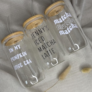 Könnte beinhalten: Drei transparente Glasbecher mit Bambusdeckeln und durchsichtigen Strohhalmen. Jeder Becher hat weißen Text: "In My Pumpkin Spice Era", "Jenny's Iced Matcha Latte" und "Matcha Matcha Matcha". Die Becher stehen auf einem neutralen Stoff.