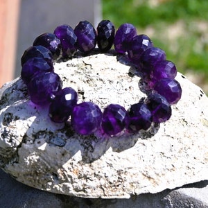 May include: A bracelet made of faceted amethyst beads. The deep purple beads are strung together to form a circular shape. The beads have a polished, reflective surface. The bracelet is displayed on a white, textured surface.