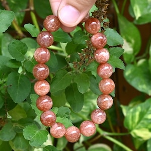 Puede incluir: Un brazalete hecho de cuentas redondas de piedra solar de color naranja rojizo. Las cuentas tienen una apariencia moteada y están ensartadas. El brazalete se muestra sobre un fondo de hojas verdes.