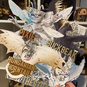 May include: A collection of paper cutouts of magical creatures from the Harry Potter universe. The creatures include a pixie, a Hungarian Horntail dragon, a Thestral, and Buckbeak the hippogriff. The cutouts are arranged on a bookshelf with a newspaper background.