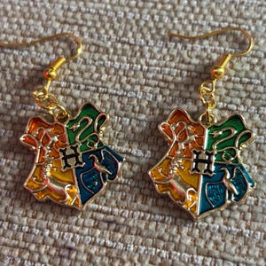 May include: A pair of gold-tone earrings featuring the Hogwarts house crest. The crest is made of enamel and features the colours red, yellow, green, and blue.