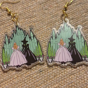 May include: A pair of clear acrylic earrings with gold-coloured hooks. Each earring features a green castle design with a pink-dressed character and a black-dressed character. The earrings are designed with a fantasy theme.