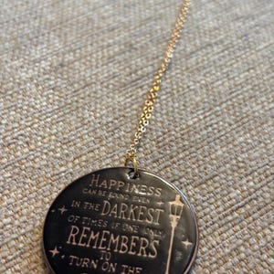 May include: A gold-colored necklace with a circular pendant. The pendant is dark with the words "HAPPINESS CAN BE FOUND EVEN IN THE DARKEST OF TIMES IF ONE ONLY REMEMBERS TO TURN ON THE LIGHT" and a street lamp design.
