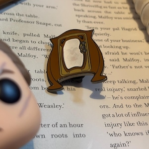 May include: A brown and gold enamel pin depicting a vintage television set with a white screen. The pin is displayed on a book with text about Harry Potter.