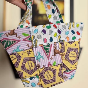 May include: A small patchwork tote bag with various patterned fabrics. The bag features a white fabric with colourful jelly bean designs, and other sections with frog and star motifs. The bag has two handles and is held by a hand.