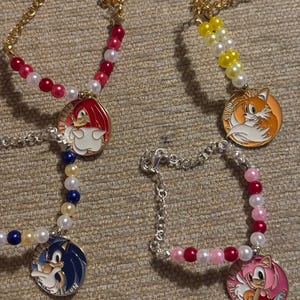 May include: Four beaded bracelets with charms featuring characters from the Sonic the Hedgehog video game franchise. The charms are round and feature Sonic, Tails, Knuckles, and Amy Rose. The bracelets are made with gold and silver chains and colourful beads.
