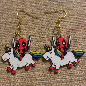 May include: A pair of novelty earrings featuring a cartoon Deadpool character riding a white unicorn. The earrings have gold-colored hooks and are set against a textured, tan background. The unicorn has a rainbow mane and tail.
