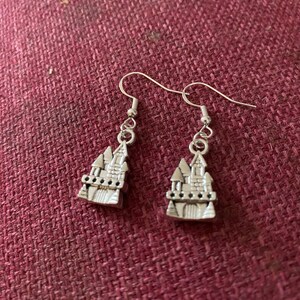 May include: A pair of silver dangle earrings featuring a detailed castle charm. The castle charm has a pointed roof, windows, and a decorative base.