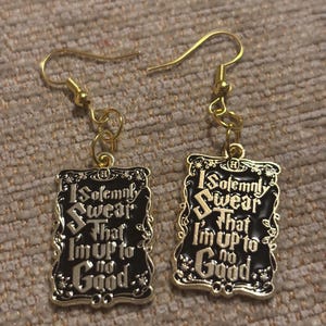 Peut inclure: Paire de boucles d'oreilles dorées avec des pendentifs rectangulaires noirs. Chaque pendentif porte l'inscription blanche "I Solemnly Swear That I'm Up To No Good". Fermeture à crochet.