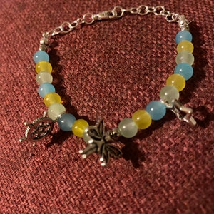 May include: A silver bracelet featuring blue and yellow beads, adorned with three silver charms: a ship's wheel, a butterfly, and a cross.