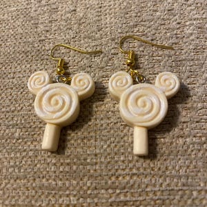 May include: A pair of gold dangle earrings with white lollipop charms. The lollipops have a swirl design and are shaped like Mickey Mouse ears.
