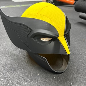 May include: A 3D printed helmet featuring a matte black finish with a textured yellow crown section. The helmet has prominent pointed side fins, a central yellow ridge, and white mesh eye coverings. The design includes a wide open chin area.