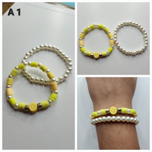 May include: Two bracelets, one with yellow and orange beads and a lemon charm, and the other with white pearl beads. The bracelets are stacked on a wrist.