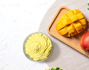 May include: A white bowl filled with yellow mango mousse, a wooden tray with a sliced mango and a whole mango, and a green leaf on a white background.