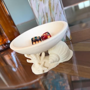 Könnte beinhalten: Eine kleine, cremefarbene Keramikschale in Form eines flachen Gefäßes, die auf einem Sockel mit skulptiertem Muschel-, Seestern- und Korallendesign ruht. Zwei sechsseitige Würfel, einer dunkelblau und einer rot, liegen in der Schale.