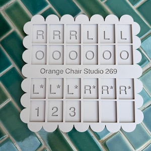 May include: A light gray rectangular organizer with a scalloped edge featuring four rows of six square compartments. The top row contains letters R and L, the second row has circles, the third row has L* and R*, and the bottom row has numbers 1, 2, and 3. The text Orange Chair Studio 269 is printed in the center.