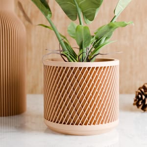 Puede incluir: Un macetero cilíndrico de color marrón claro con un patrón geométrico, que contiene una planta verde con hojas grandes. El macetero está sobre una superficie blanca.
