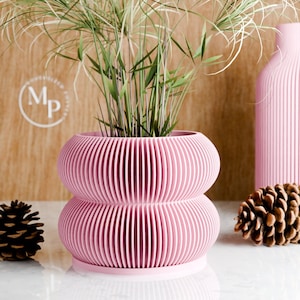 Puede incluir: Un macetero rosa de dos niveles con un diseño acanalado. El macetero está lleno de plantas verdes y se encuentra sobre una superficie de madera.