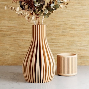 May include: A wooden vase with vertical stripes holds dried flowers and foliage. A small, matching wooden container sits beside the vase. The vase and container are made of light-colored wood and are set against a light brown background.