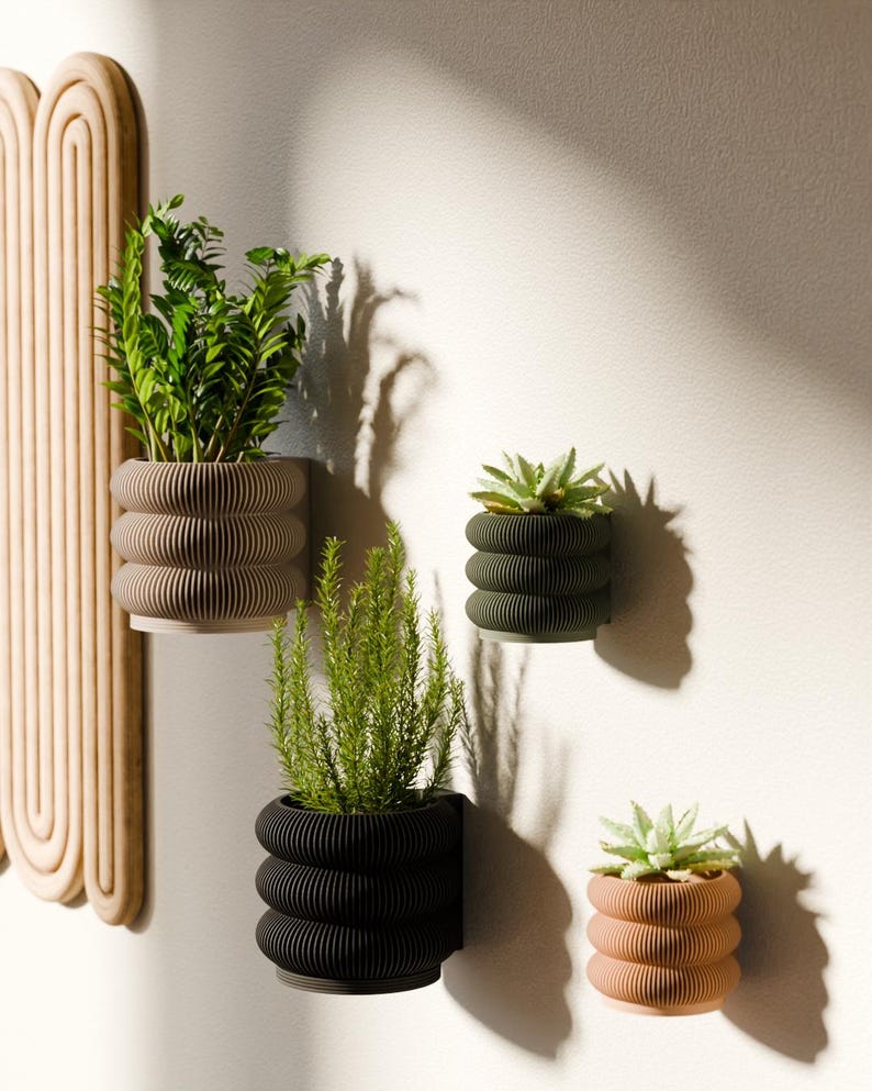 May include: A collection of wall-mounted planters in various colors, including beige, olive green, black, and peach. Each planter features a unique, ribbed design and holds different types of greenery, creating a modern and stylish display. The planters are attached to a white wall.