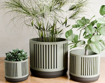 May include: Three modern, cylindrical planters in varying sizes. The planters are light green with vertical cut-out designs and dark brown bases. Each planter holds a different type of greenery, creating a stylish indoor or outdoor decor.