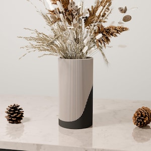 May include: A cylindrical vase with vertical striped pattern in cream and dark red, and a black curved section. The vase holds a dried floral arrangement with beige and brown tones. Two pinecones sit on the surface.