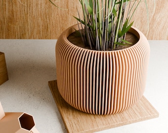 May include: A light brown, cylindrical planter with vertical grooves, made of wood, with a green plant growing inside. The planter is sitting on a light brown wooden base.