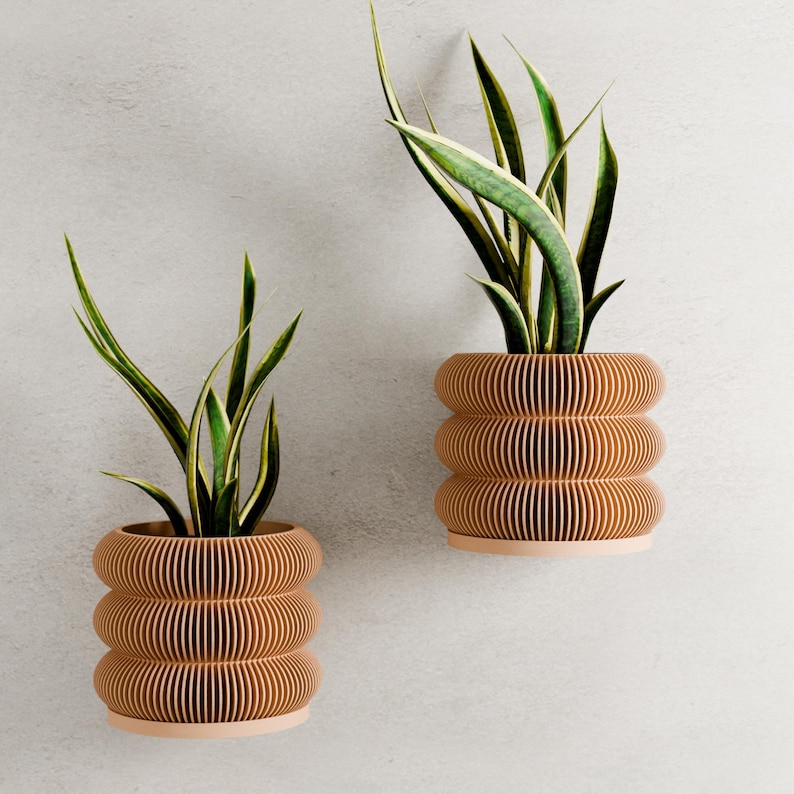 May include: Two wall-mounted, stacked, cylindrical planters with a light brown, ribbed design. Each planter holds a snake plant with green and yellow striped leaves. The planters have a light pink base and are mounted on a light gray wall.
