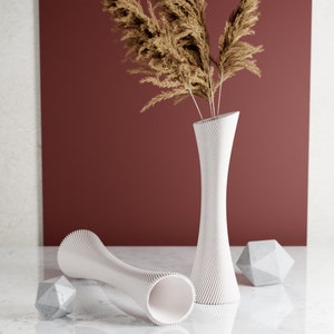 May include: Two white ceramic vases with a textured, ribbed design. The vases are empty and are sitting on a white surface. A bouquet of dried pampas grass is in the taller vase.