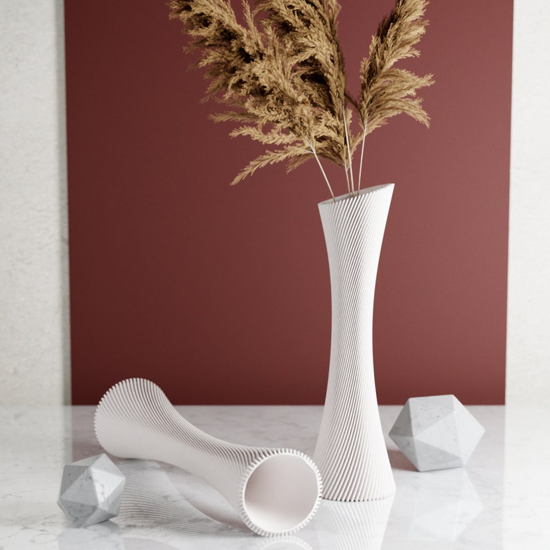 May include: Two white ceramic vases with a textured, ribbed design. The vases are empty and are sitting on a white surface. A bouquet of dried pampas grass is in the taller vase.