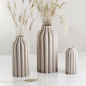 May include: Three beige ceramic vases with a ribbed design. The vases are arranged in a row on a white surface. The tallest vase is in the center, and the two smaller vases are on either side. The vases are empty.