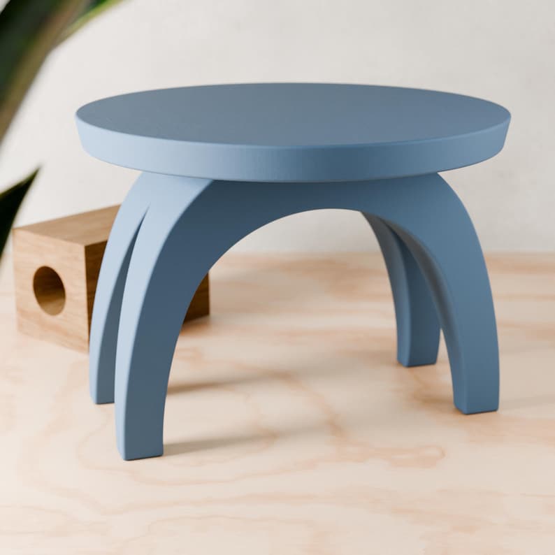 May include: A small, round, light blue wooden stool with a unique arched leg design. The stool is made of solid wood and has a smooth, painted finish.