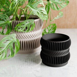 May include: A black and a beige decorative plant pot with a textured, ridged design. The black pot is in the foreground, and a larger beige pot with a plant is behind it. The plant has large green leaves with holes.