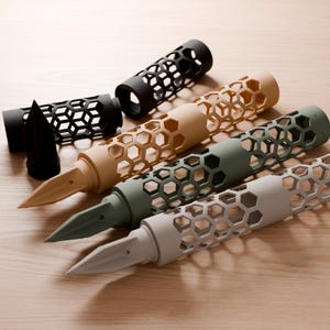 May include: A collection of four fountain pens featuring a cylindrical barrel with a cut-out honeycomb lattice pattern. The pens are available in matte black, tan, olive green, and light gray, each with a matching nib and a separate cap.