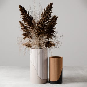 May include: Two decorative vases. The larger vase is white with vertical lines and a curved beige accent. The smaller vase is brown with vertical lines and a black accent. Both vases are cylindrical. The larger vase contains dried brown and beige grasses.