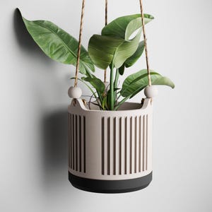 Puede incluir: Un macetero colgante con un cuerpo cilíndrico beige claro con líneas verticales recortadas. El macetero tiene una base negra y está suspendido por una cuerda marrón. Contiene una planta verde y frondosa, creando una estética moderna.