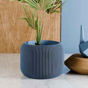 May include: A blue planter with vertical grooves, made of a ribbed material, holds a green potted plant with long leaves. The planter is sitting on a light gray surface.