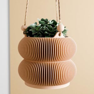 May include: A light tan hanging planter with a ribbed, double-bulbous shape. The planter is suspended by thin twine cords featuring two round wooden beads at the top attachment points. A small green succulent plant is potted inside the container.